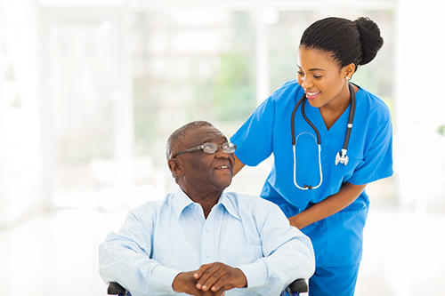 Nurse in blue scrubs smiling at elderly man sitting in wheelchair next to her